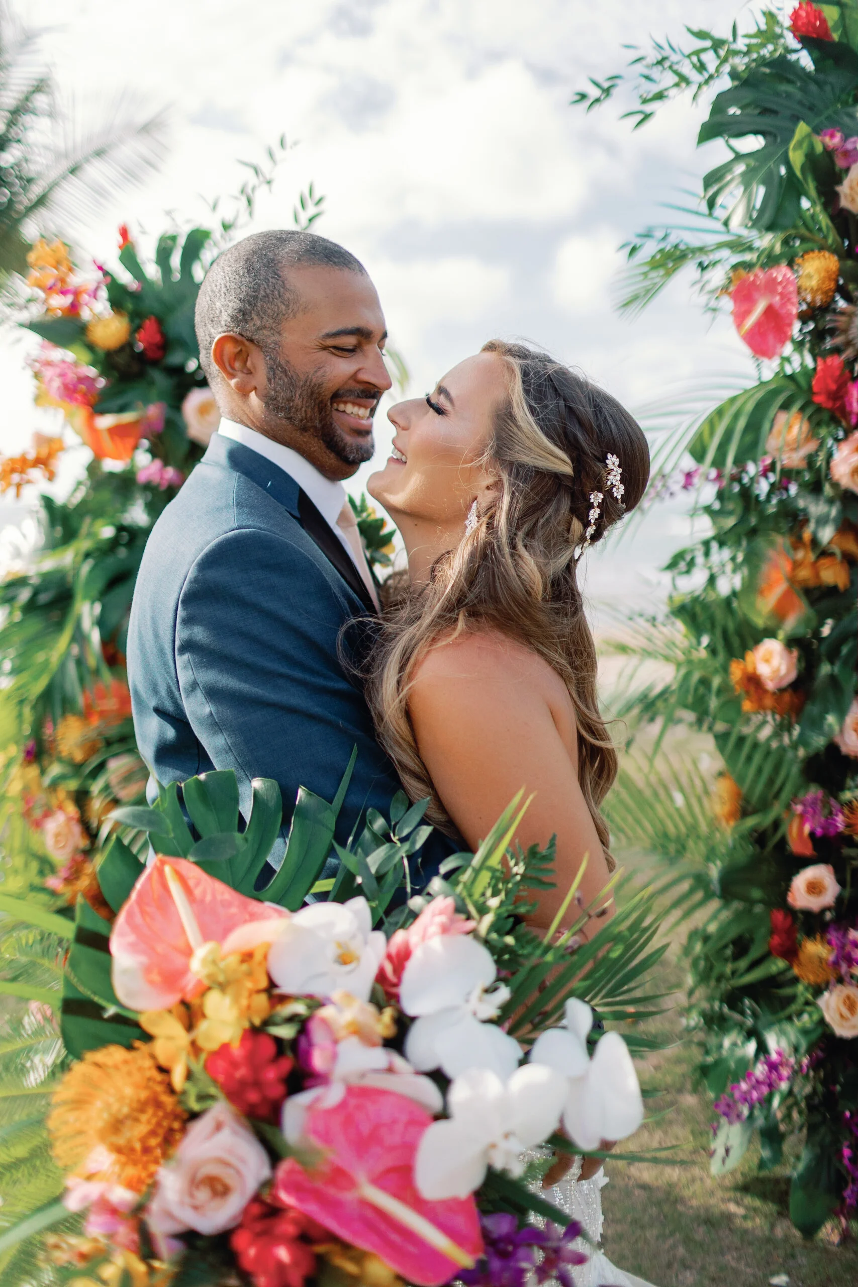 Couple embracing with vibrant floral arch featuring anthuriums, orchids, and lush greenery at Loulu Palms, Oʻahu wedding. — Wedding Event Oahu Hawaii Florals Flowers Florist Creative Concepts by Jeff Alencastre