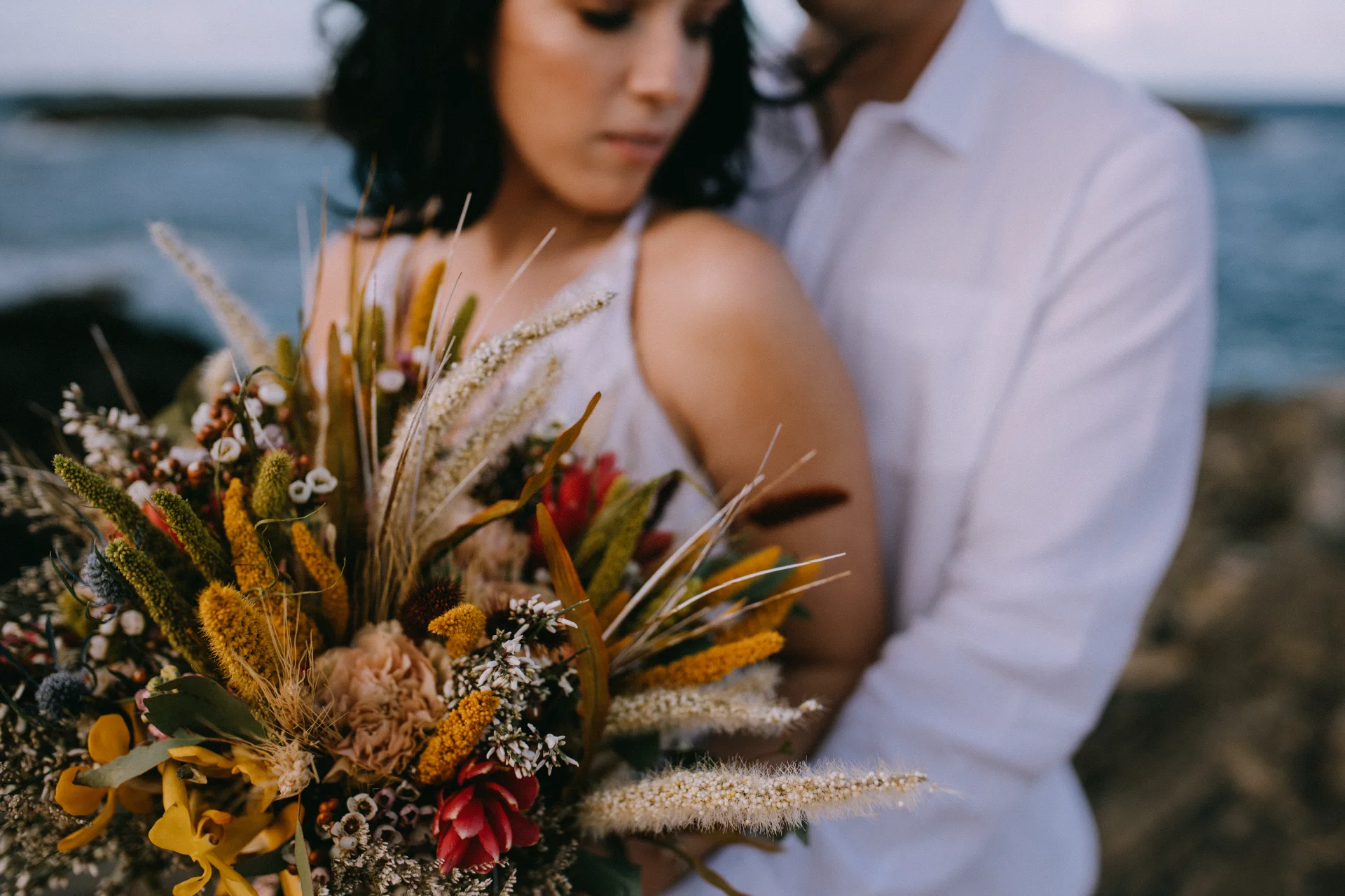 bride holding rustic bouquet with protea and wildflowers by ocean on Oʻahu