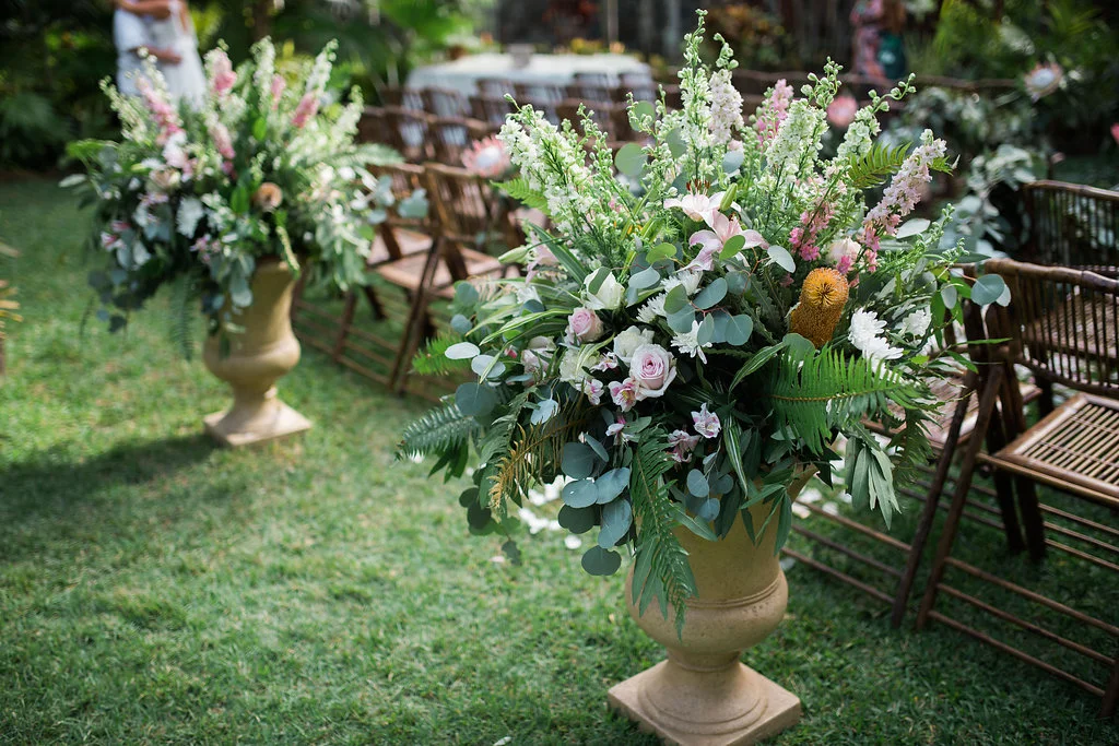 Elegant outdoor wedding florals on Oʻahu featuring lush arrangements of pink roses, white lilies, green ferns in brown urns