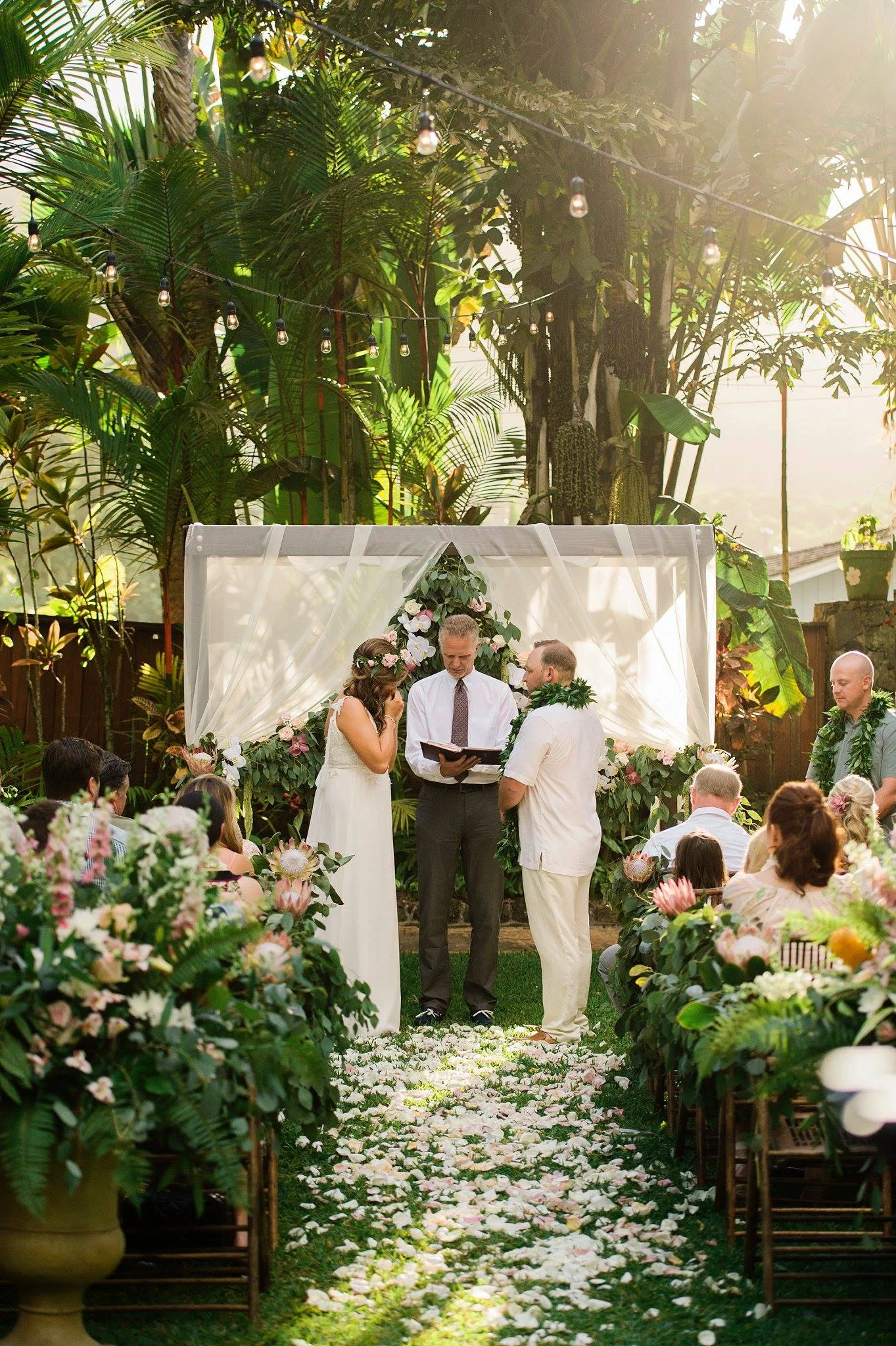 tropical outdoor wedding ceremony on Oʻahu with lush floral arch, pink proteas, and greenery surrounding couple and officiant