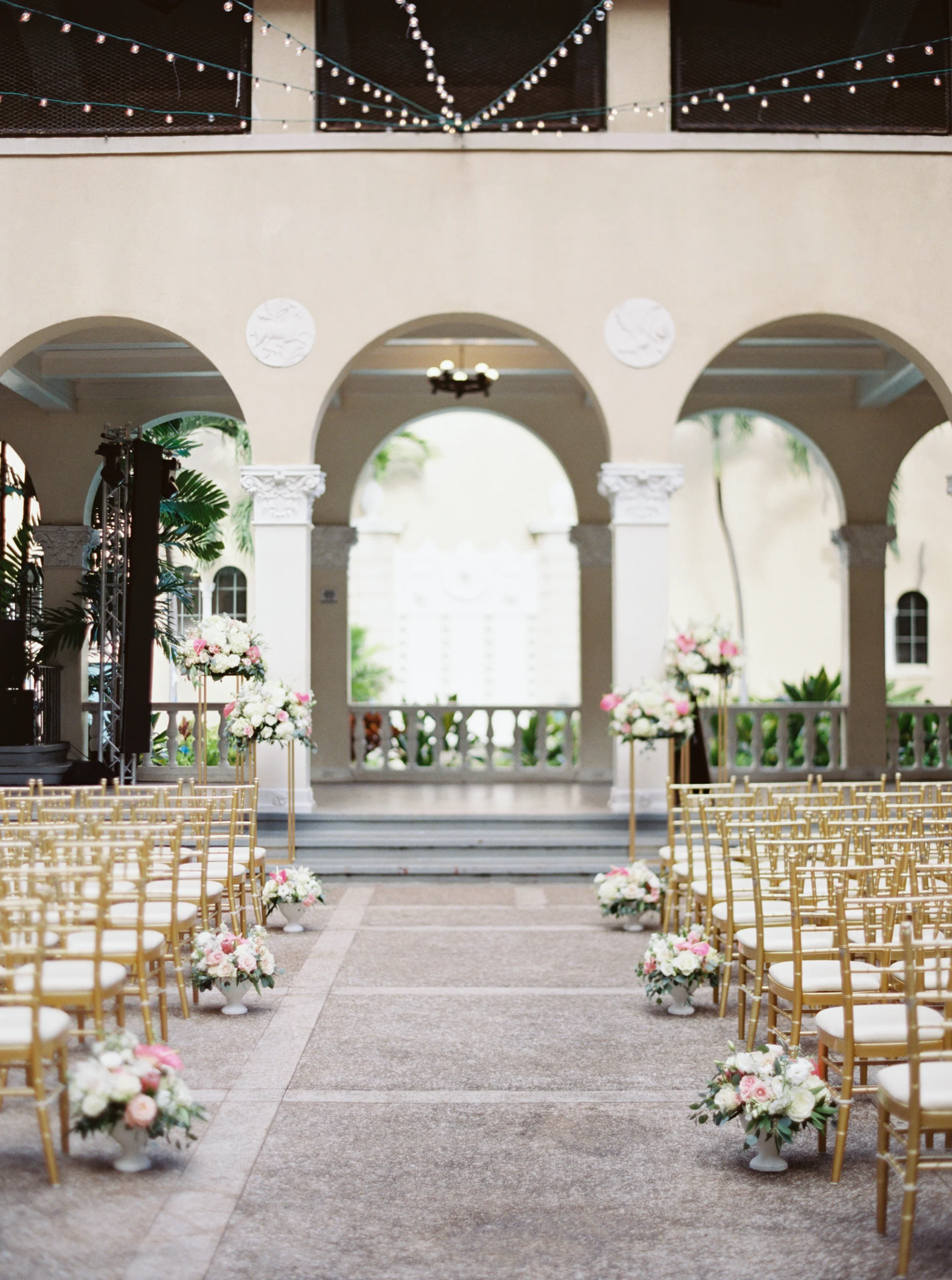 Elegant wedding ceremony setup at Cafe Julia, Oʻahu, featuring gold chairs, pink and white floral arrangements, and string lights.