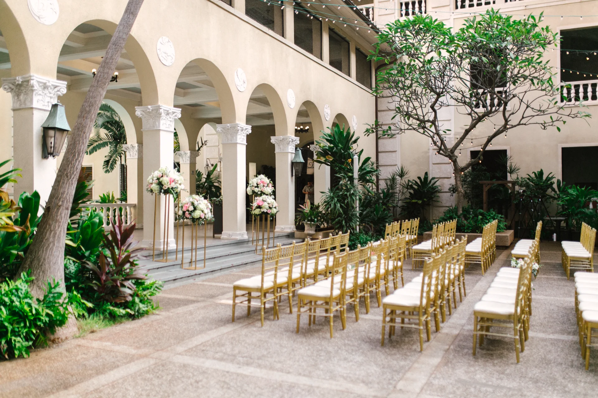 Elegant wedding setup at Cafe Julia, Oʻahu, with gold chairs and luxurious floral arrangements in a historic courtyard.