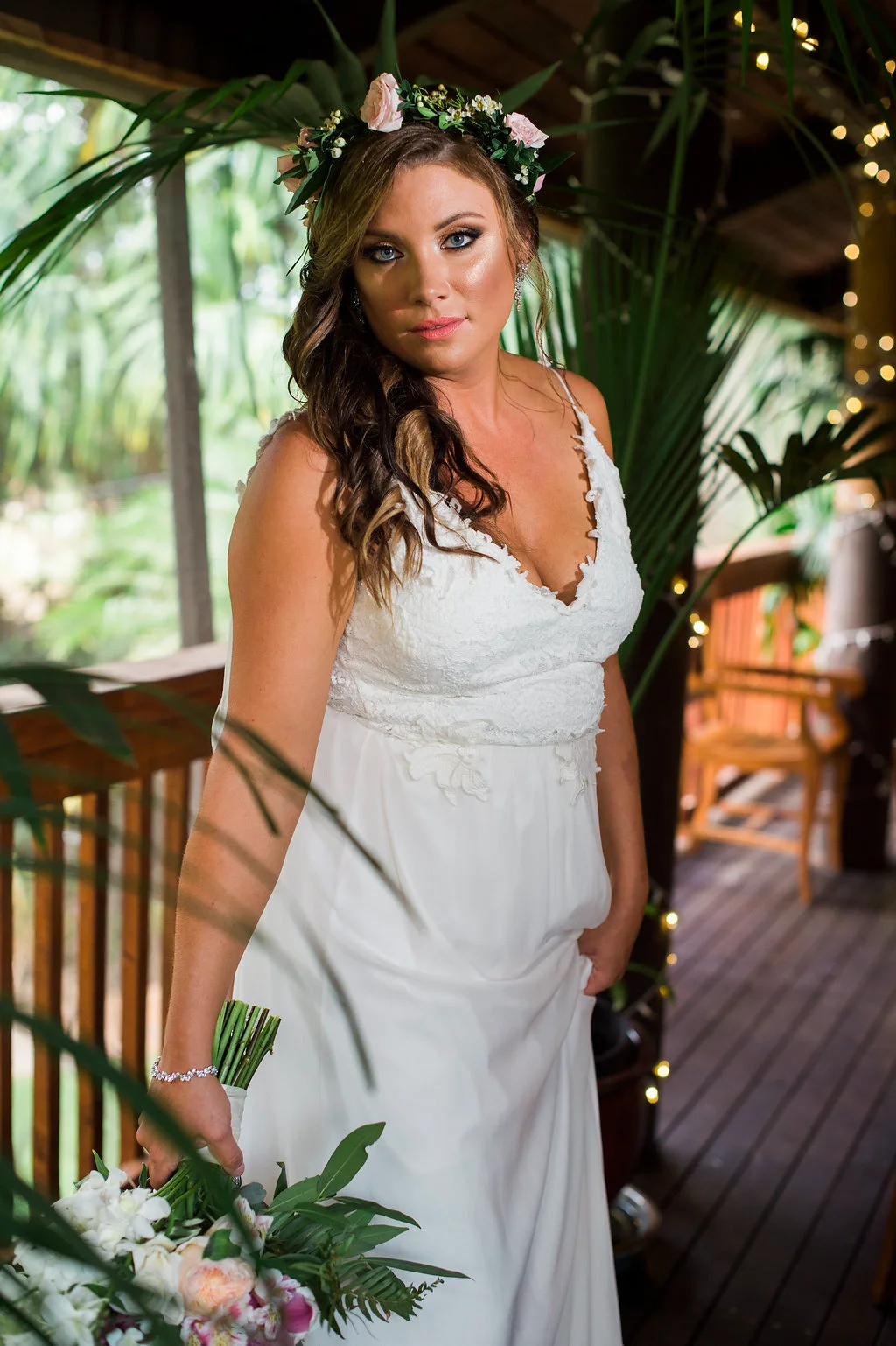 Bride in white gown holding bouquet with peonies and greenery, wearing flower crown, surrounded by tropical foliage on Oʻahu.