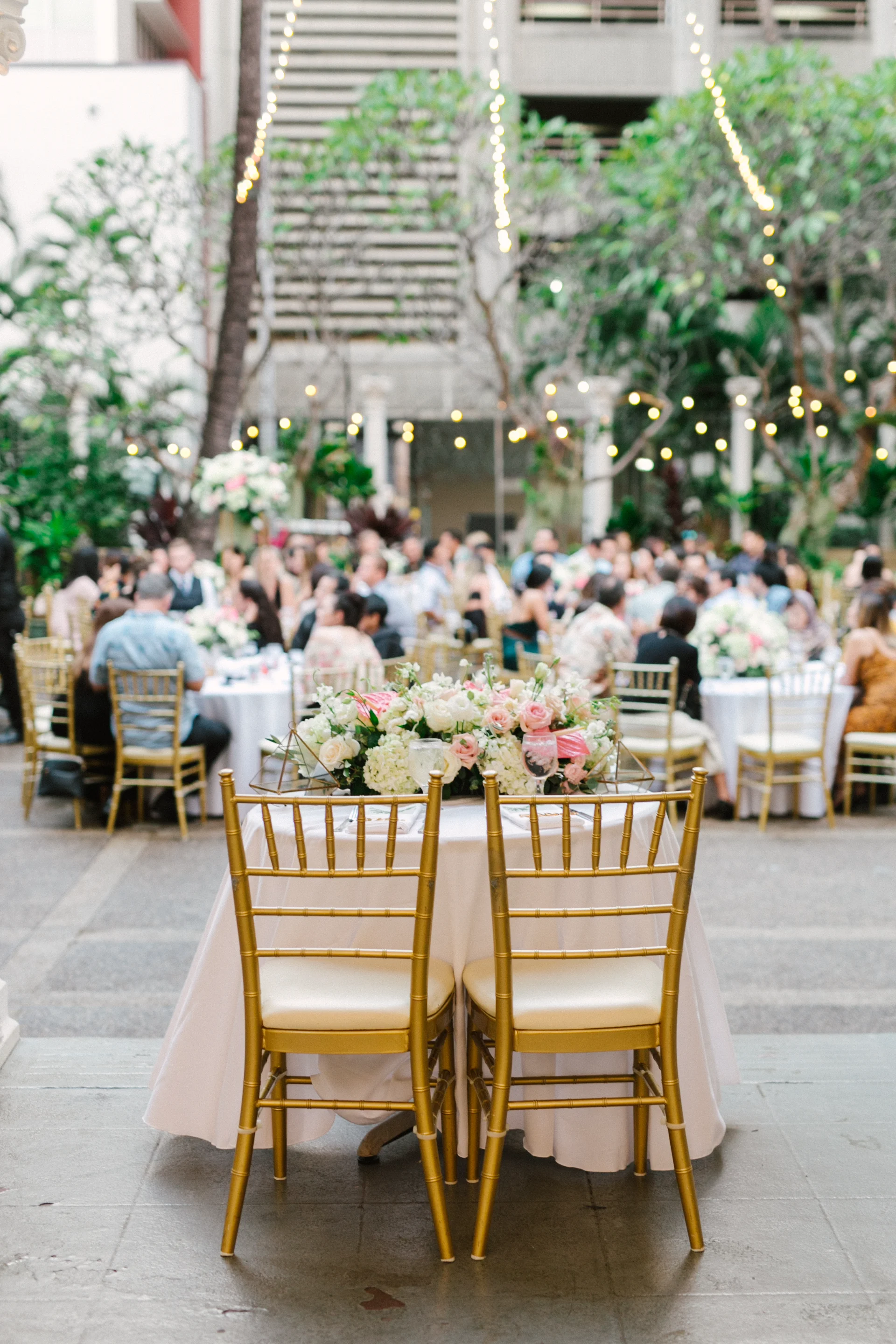 Elegant outdoor wedding reception at Cafe Julia, Oʻahu, featuring gold chairs and tables adorned with white and pink floral arrangements.