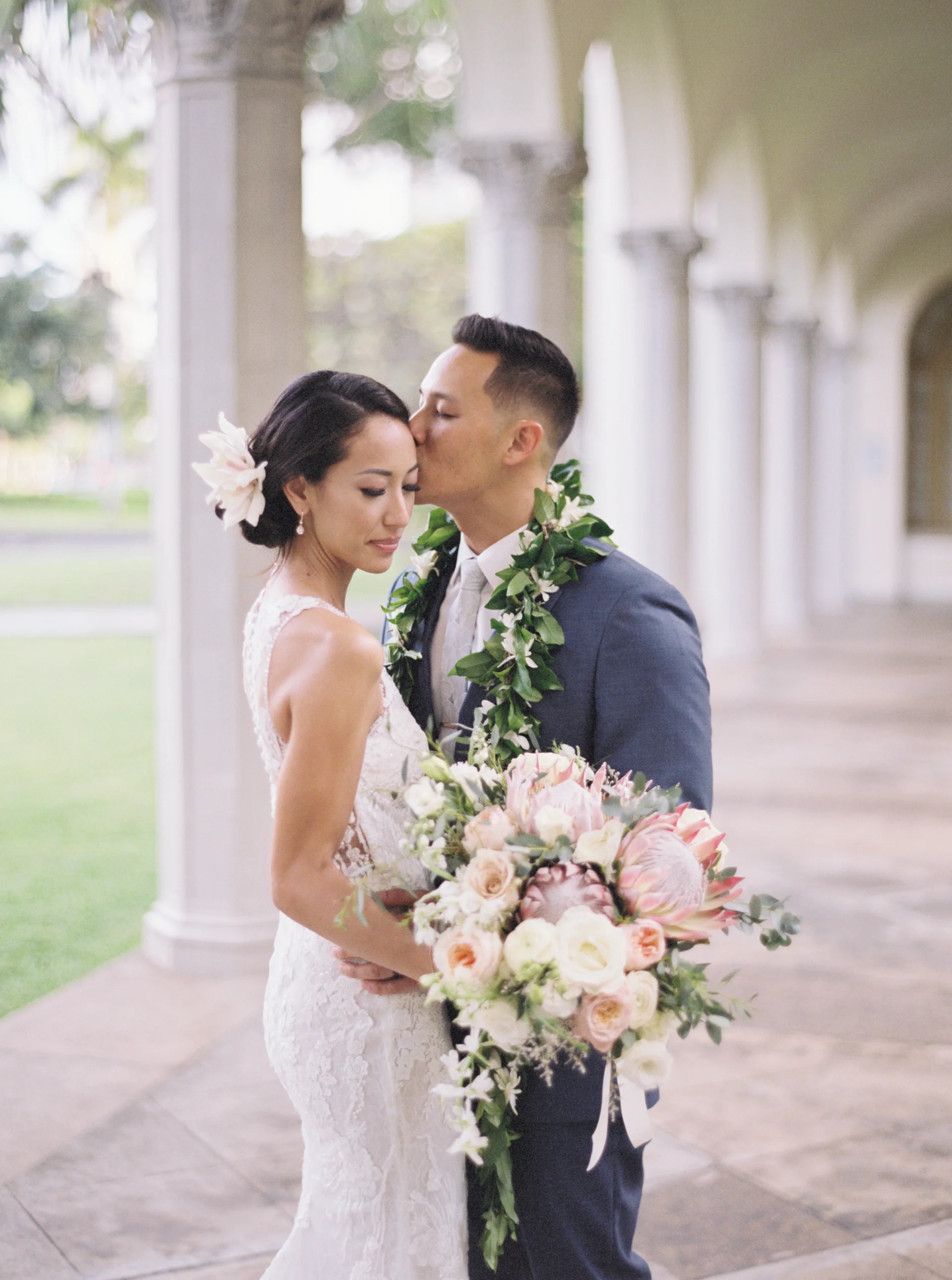Bride and groom at Cafe Julia, Oʻahu with a bouquet of roses and protea, wearing leis in an elegant wedding setting.
