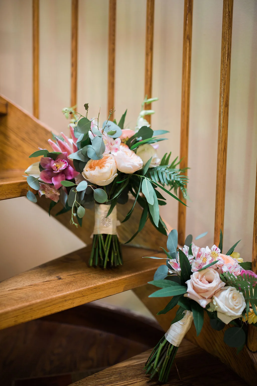Elegant wedding bouquets with pink orchids, peach roses, and eucalyptus displayed on wooden stairs in Oʻahu venue.
