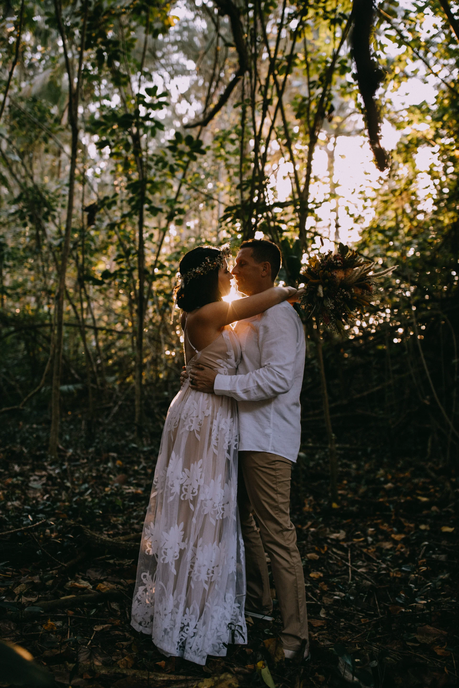 Couple embracing in forest with sunset backlight, bride holding rustic bouquet, wearing floral lace dress, Oʻahu setting.