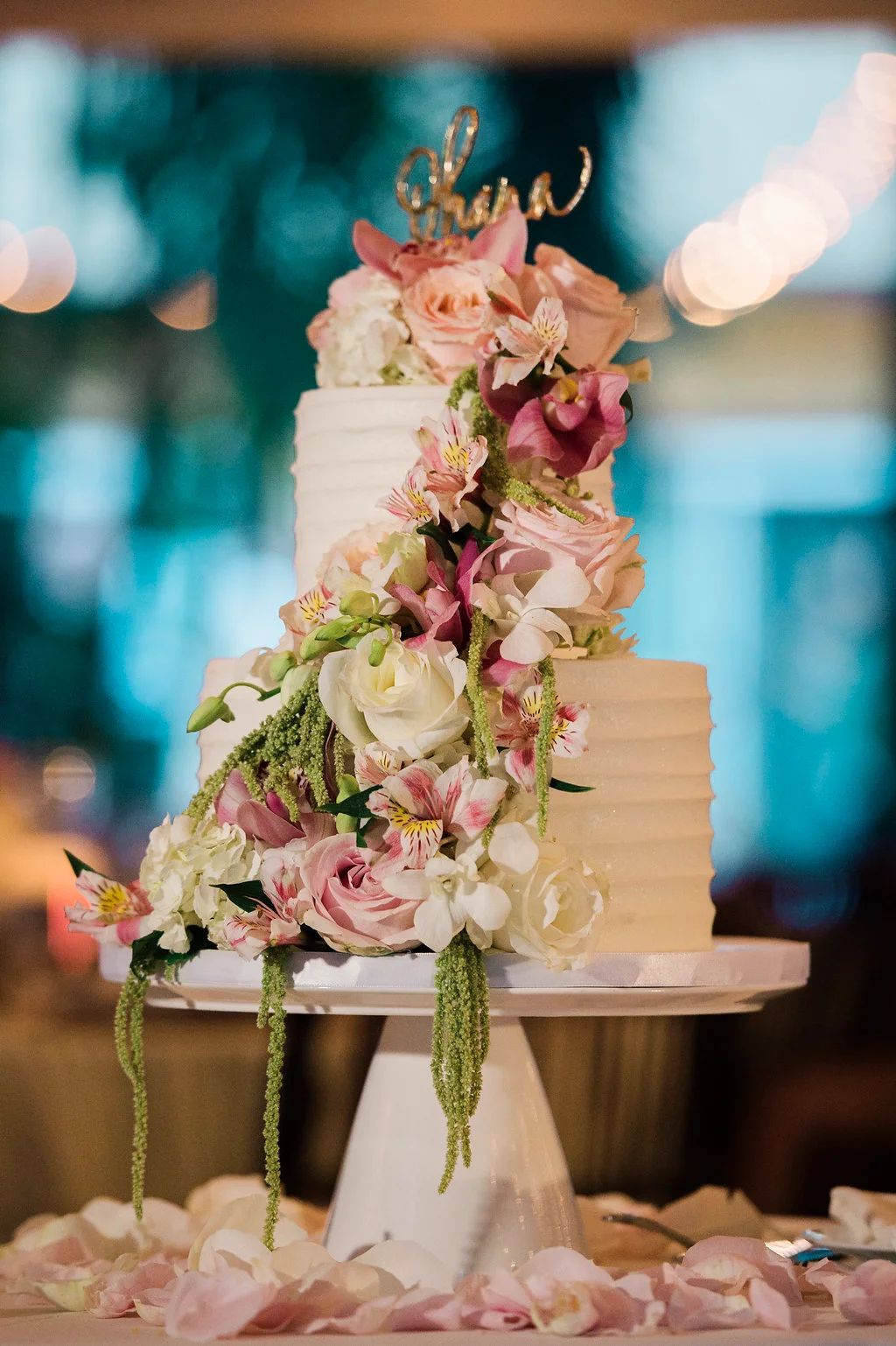 Elegant wedding cake adorned with pink and white roses, orchids, and cascading greenery, set in a luxurious Oʻahu venue.