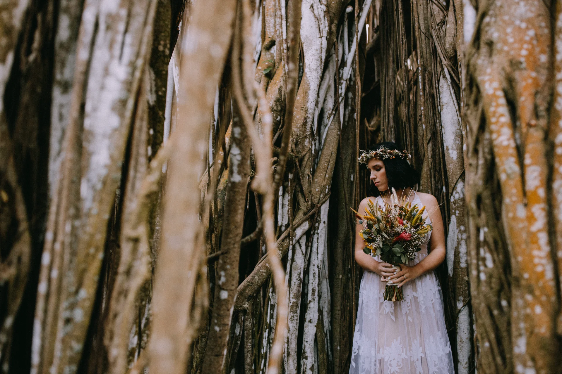 Bride with floral bouquet and crown in forest setting, surrounded by tall banyan tree roots on Oʻahu, Hawaii.