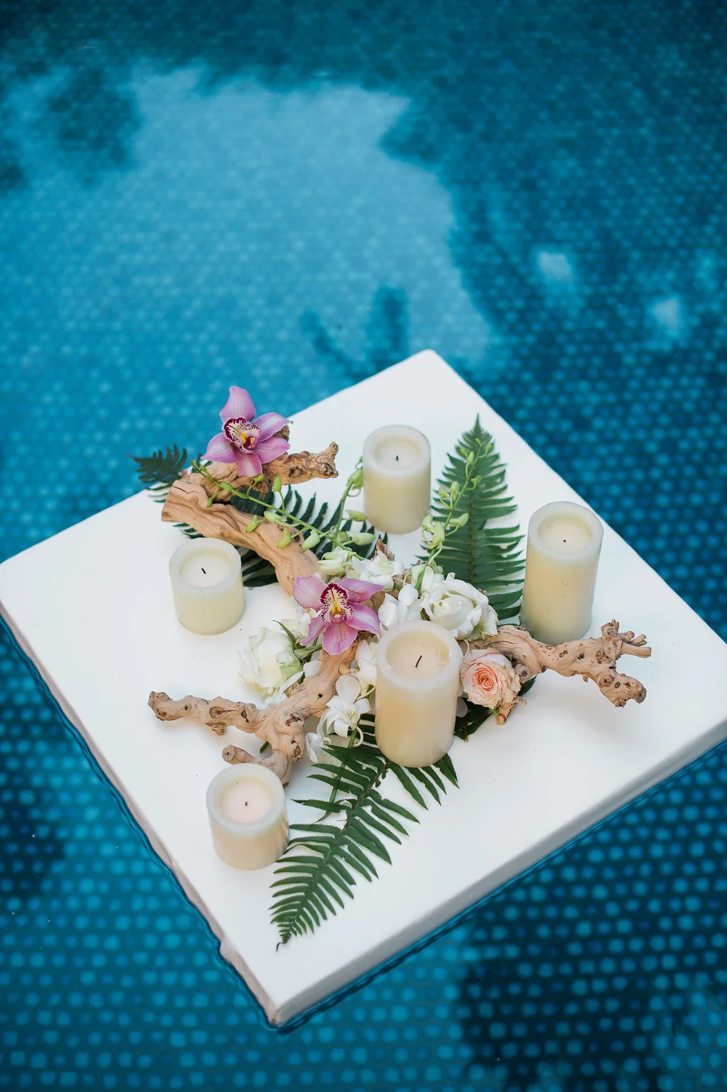 Tropical wedding centerpiece with pink orchids, white roses, ferns, driftwood, and candles on a floating table over a pool in Hawaii.