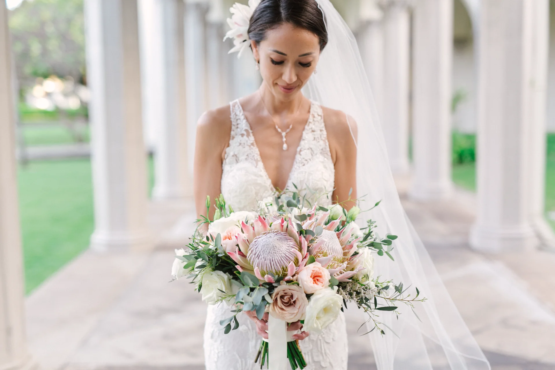 Bride at Cafe Julia holding a bouquet with proteas, roses, and eucalyptus, wearing a lace gown and veil, Oʻahu wedding