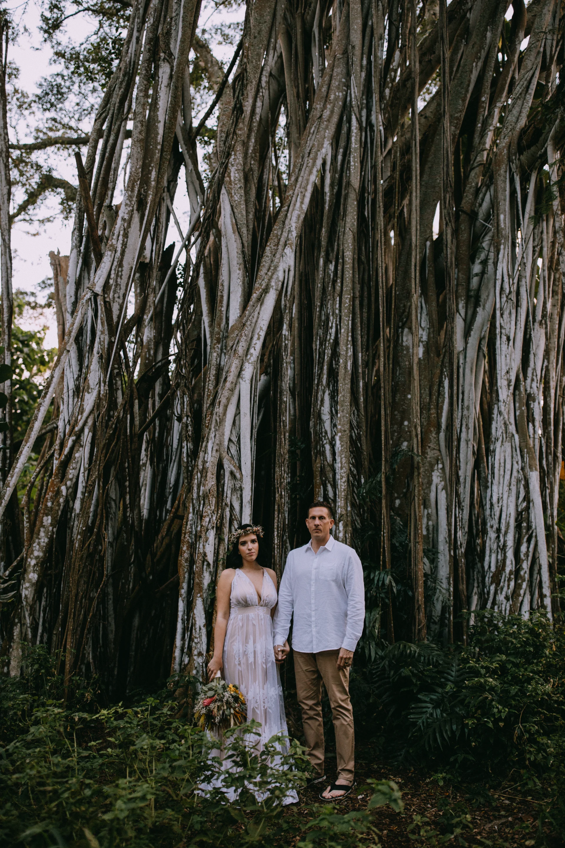 Couple in wedding attire with floral bouquet standing by a towering banyan tree on Oʻahu.