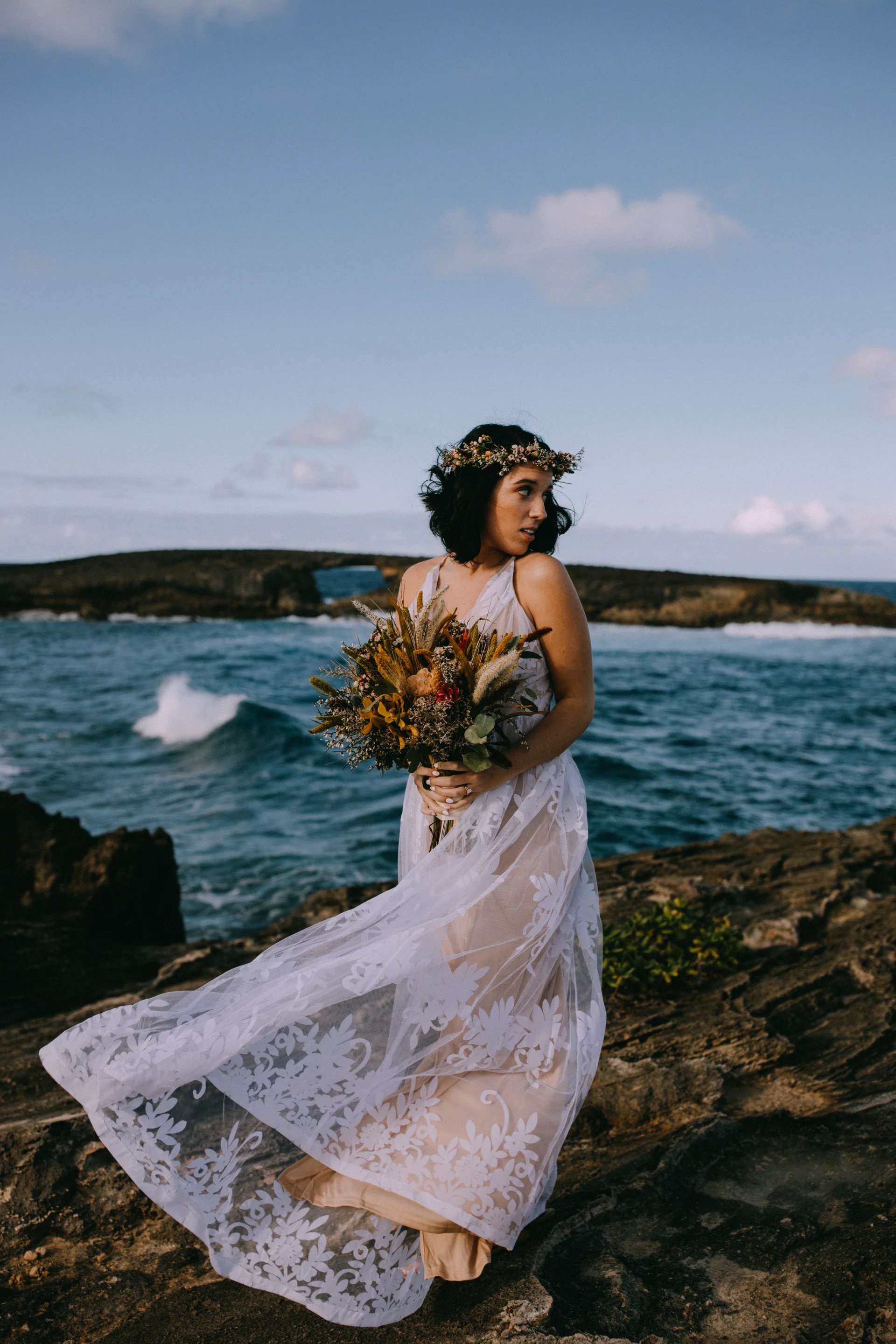Bride in lace dress holds rustic bouquet on rocky Oʻahu beach with ocean waves and floral crown.