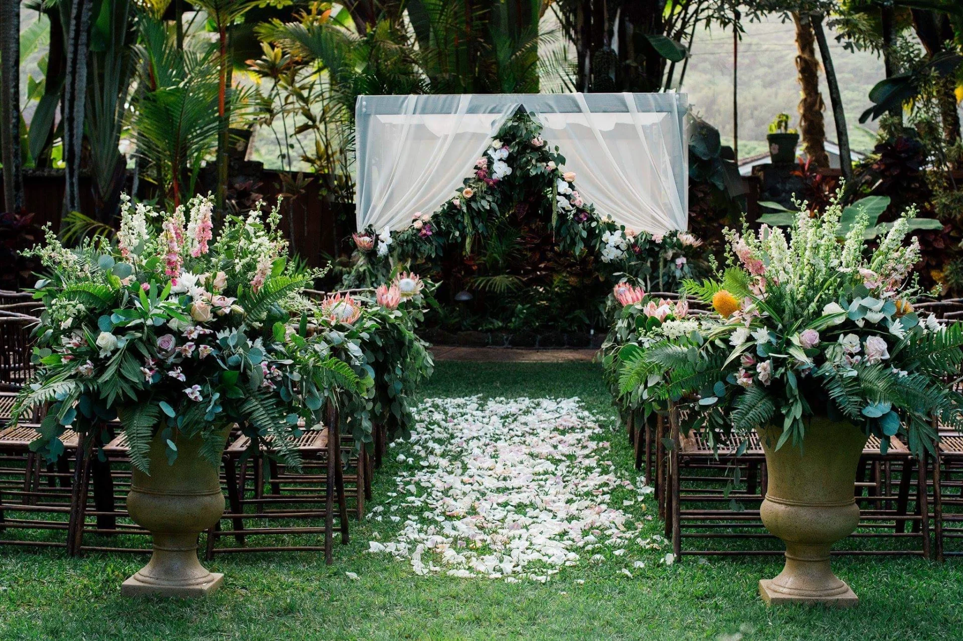 Tropical wedding ceremony setup on Oʻahu with lush greenery, white rose petals, pink and white flowers, and draped arch.