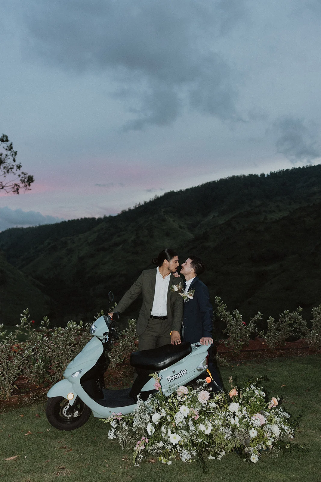 Couple in wedding attire with a scooter, surrounded by lush floral arrangements and greenery, set against an Oʻahu mountain backdrop at dusk.