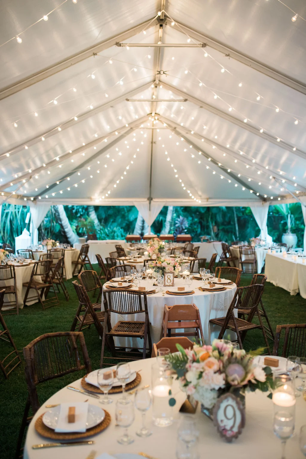 Elegant wedding reception setup under a white tent with string lights in Oʻahu, featuring floral centerpieces with pink and white flowers.