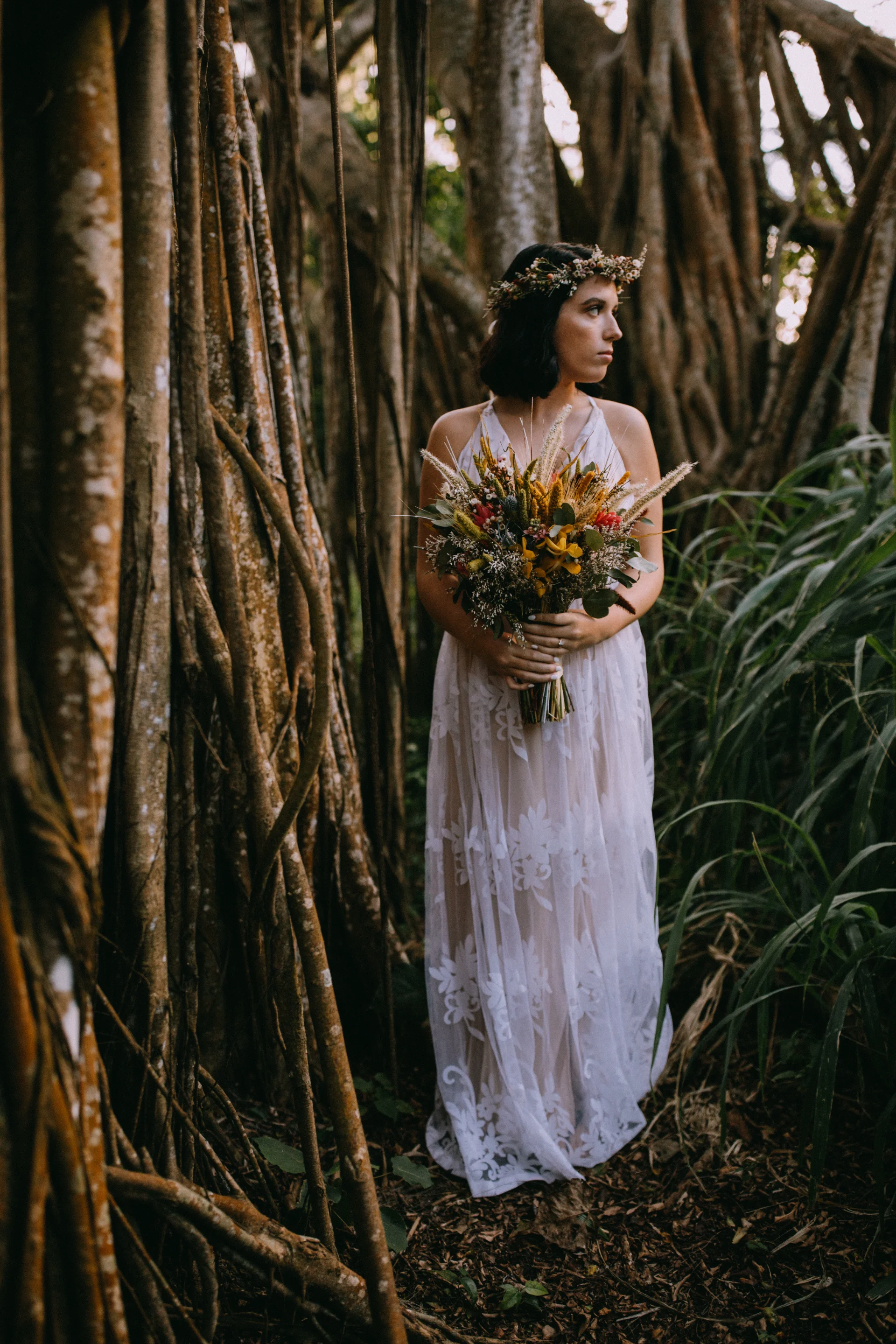 Bride in lace dress holding dried floral bouquet stands amidst tropical roots and foliage on Oʻahu
