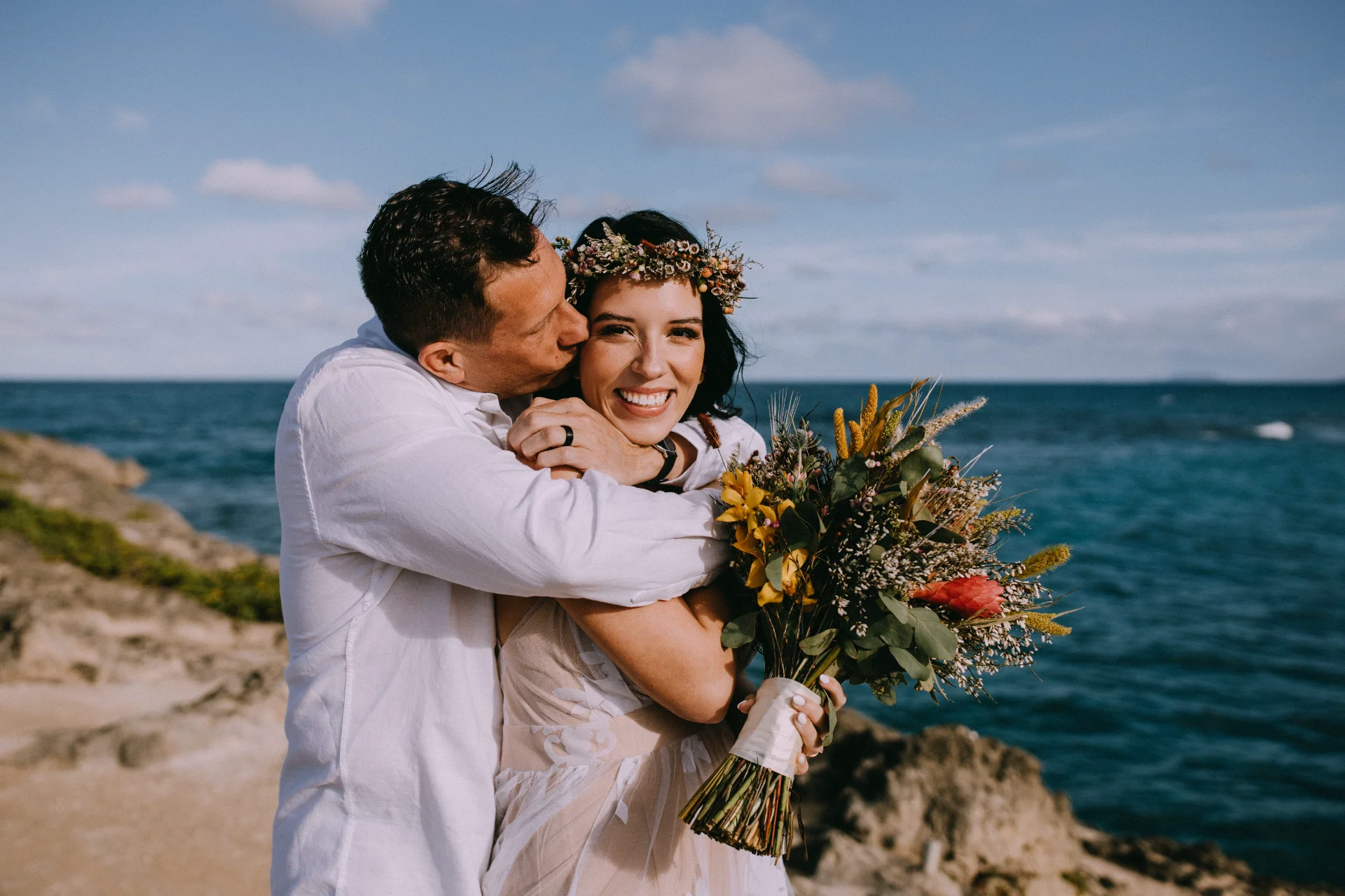 Bride holding a bouquet with tropical flowers and a floral crown, embraced by groom on a rocky Oʻahu beach.