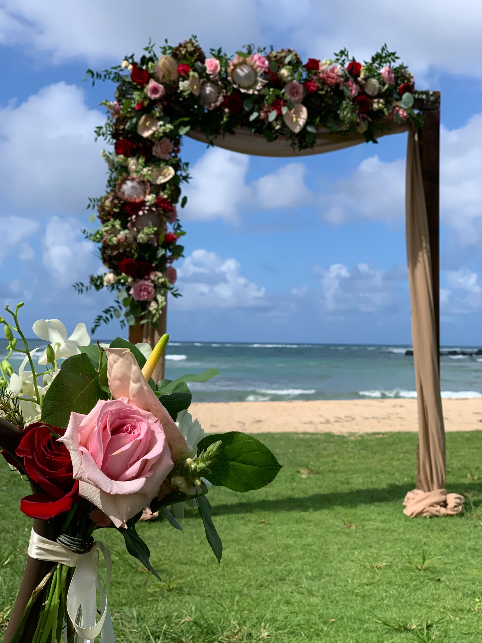 Beachfront wedding arch on Oʻahu decorated with red and pink roses, greenery, and a bouquet of tropical flowers.