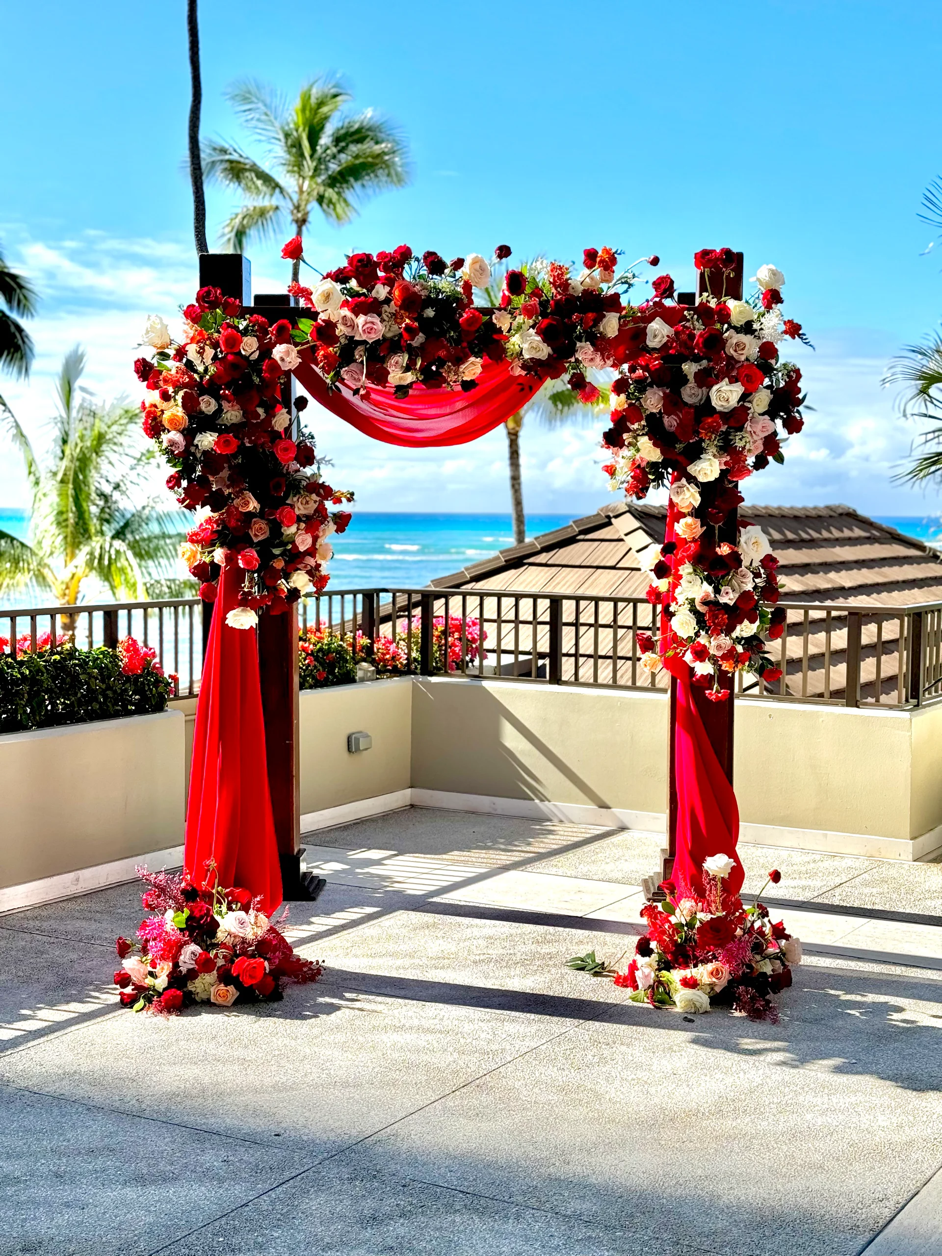 Elegant floral arch with red and white roses, draped with red fabric, set against a tropical ocean backdrop in Oʻahu, Hawaii.
