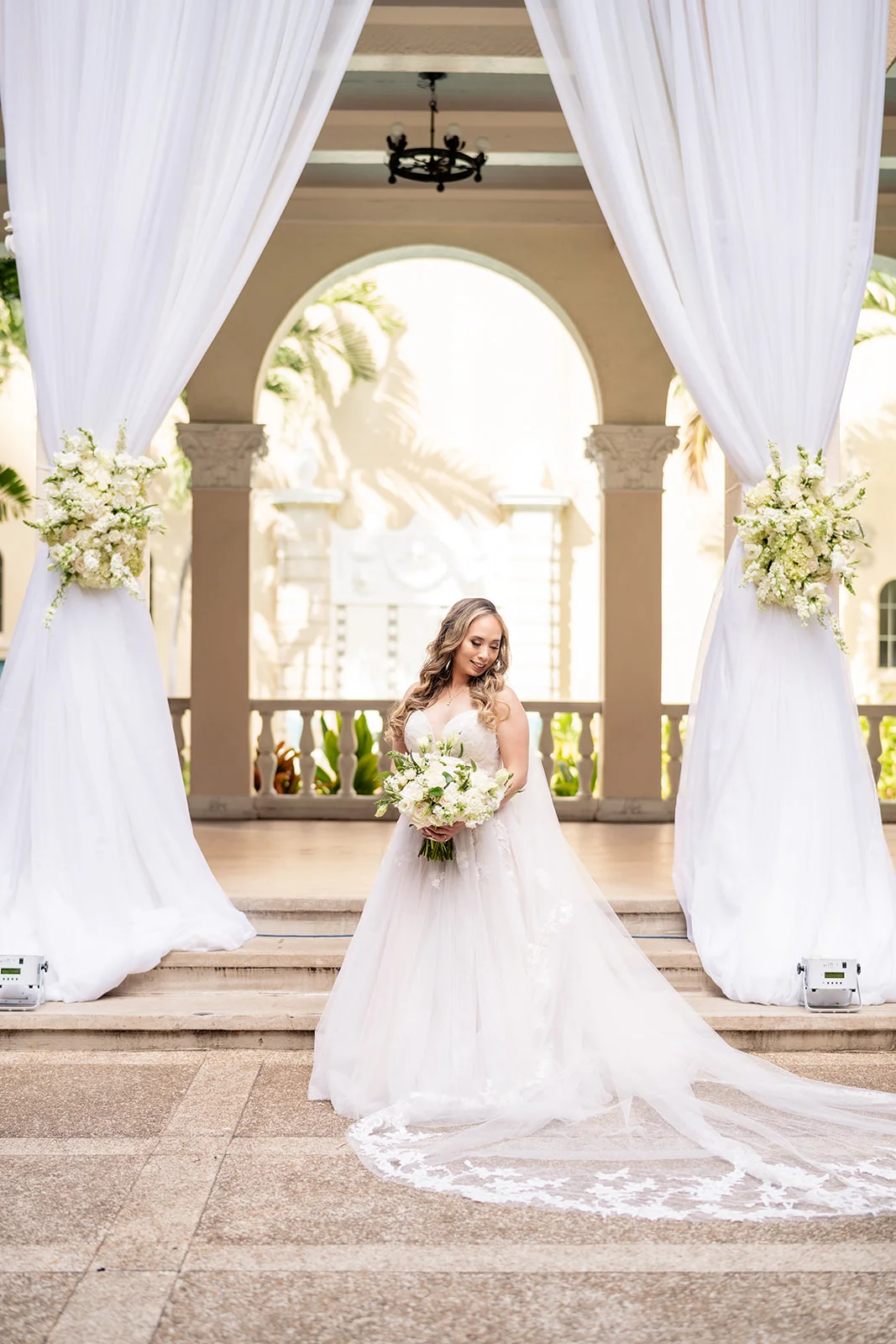 Bride holding white floral bouquet with elegant drapery and floral arrangements at Cafe Julia, Oʻahu wedding.