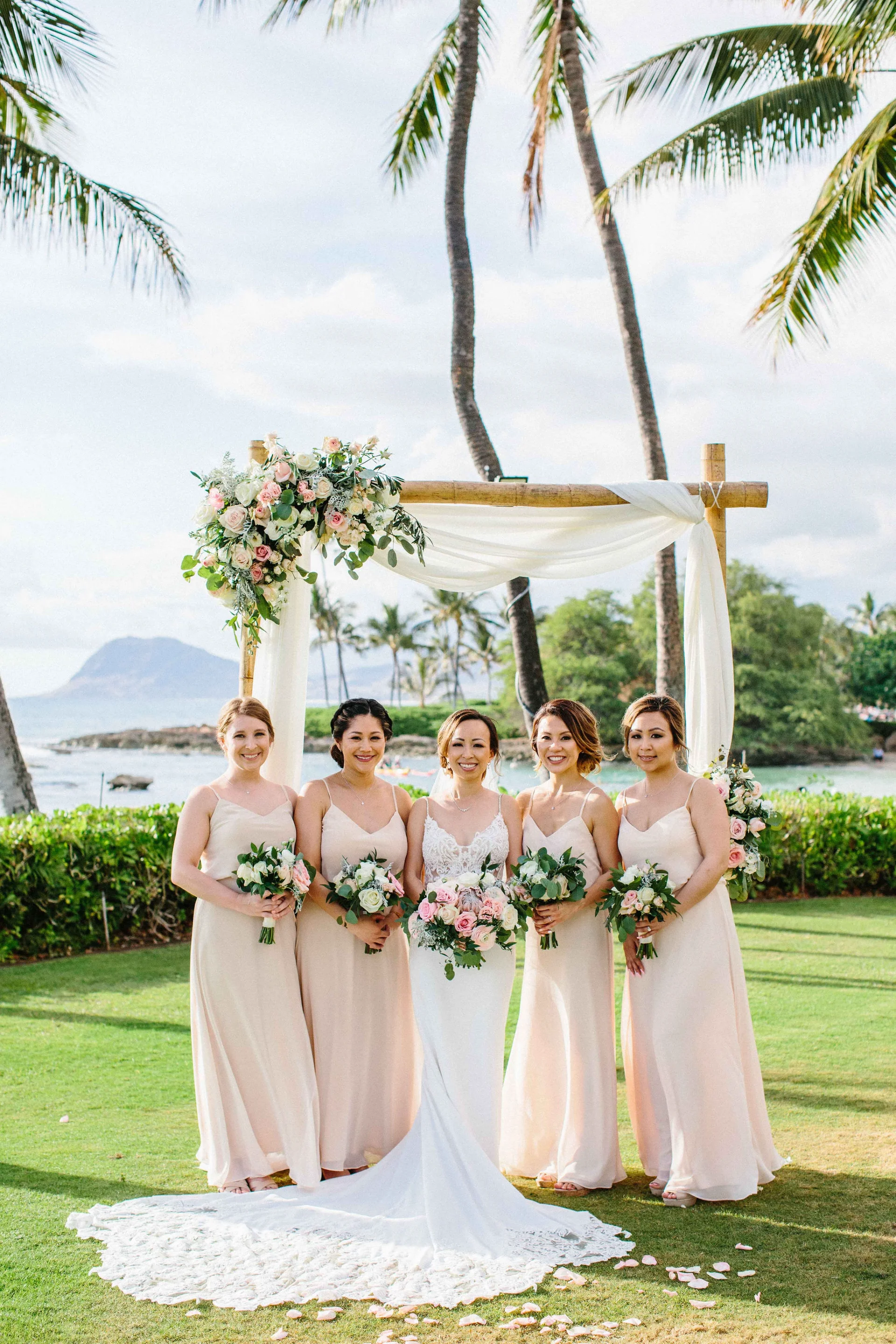 Bridal party in pale pink dresses with pink and white bouquets under floral arch at Lanikūhonua Cultural Institute, Oʻahu